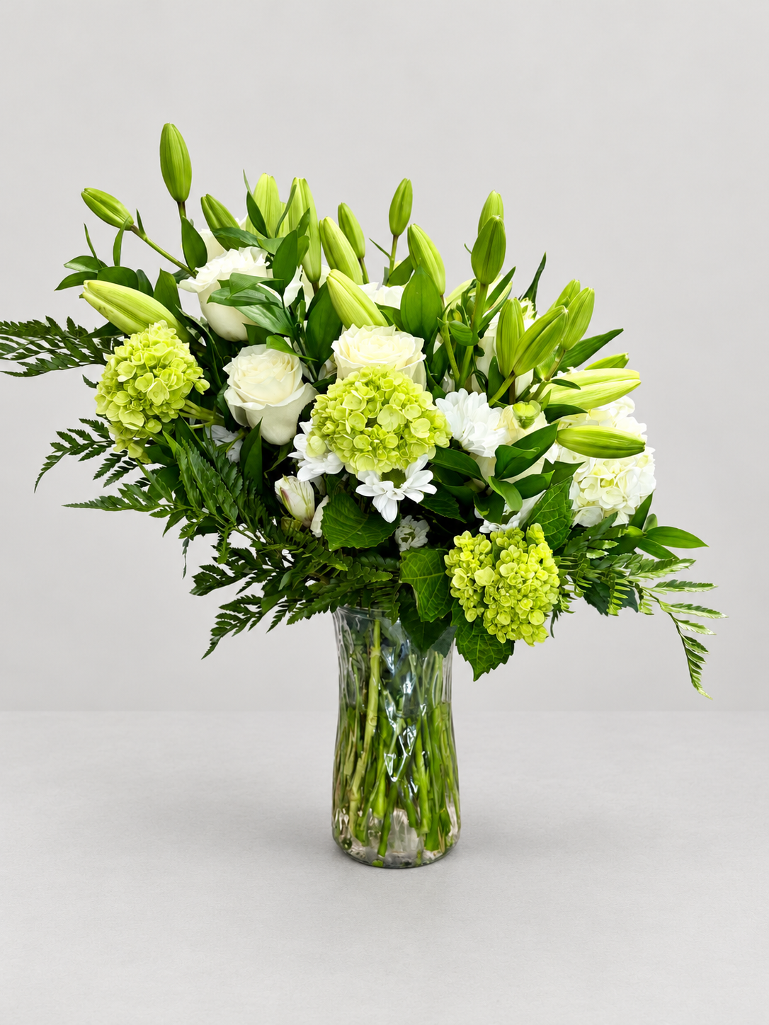 Gentle Remembrance arrangement of lilies, white roses, hydrangeas and greenery in a clear glass vase against a light grey background — Bella Rosa MTL