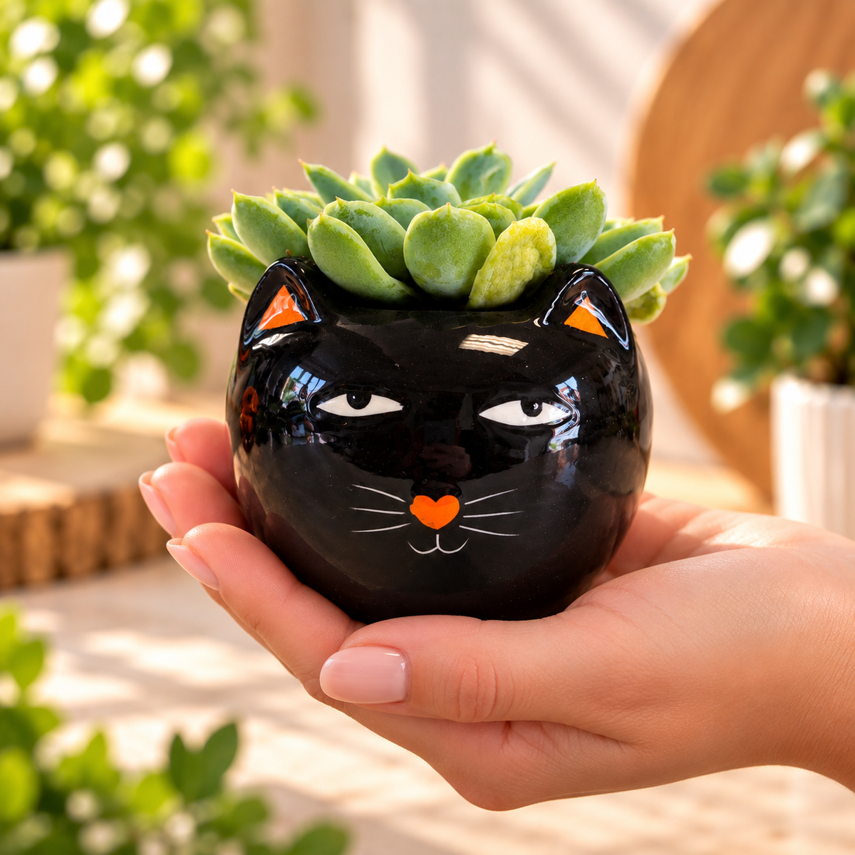 A hand holds a glossy black cat-shaped planter with a green succulent on top. The planter has white painted eyes and nose details, orange inner ears, and a small orange heart on the nose, set against a softly blurred outdoor background with plants.